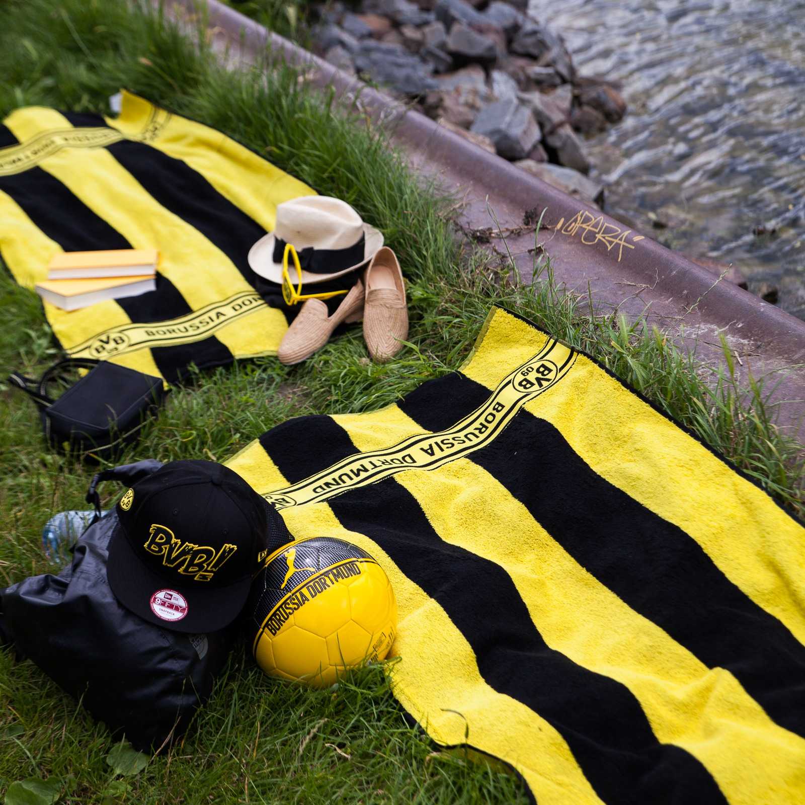 Zwei BVB-Duschtücher mit schwarzgelben Blockstreifen und BVB-Emblem in der Draufsicht am Ufer, dazu liegen BVB-Merchandise-Artikel auf dem Gras.