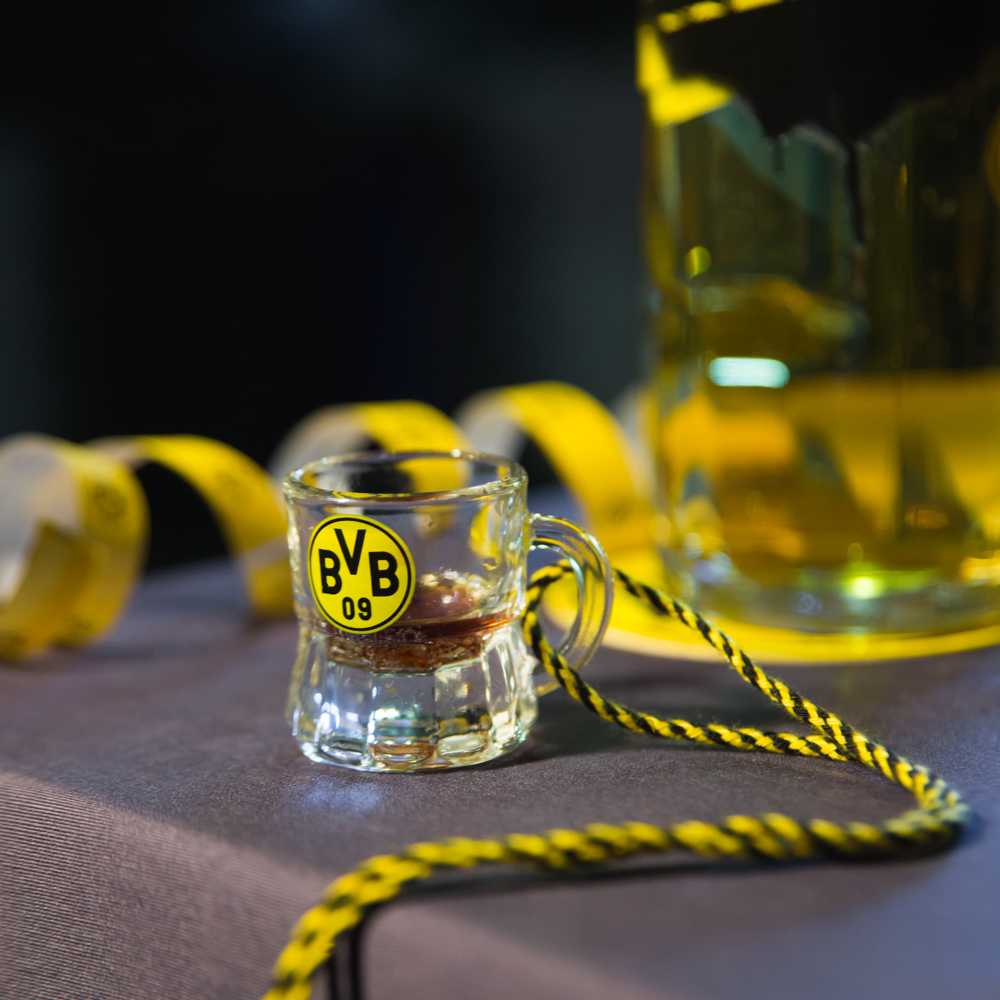 Detailansicht des BVB-Schnapsglases mit schwarzgelber Kordel und BVB-Emblem, auf einem Tisch mit unscharfer Dekoration im Hintergrund.