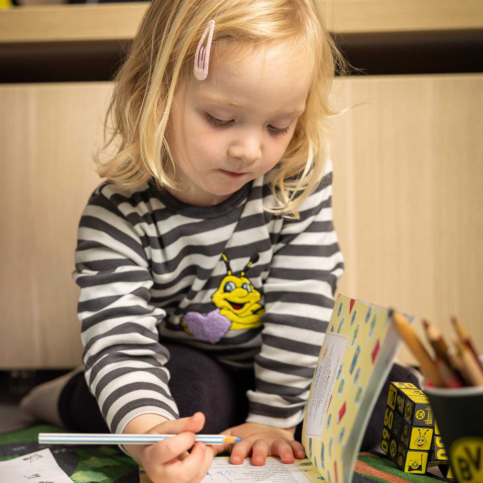 Ein Kind trägt das schwarz-weiß gestreifte BVB Longsleeve Baby mit EMMA-Motiv auf der Vorderseite, Frontalansicht beim Malen.