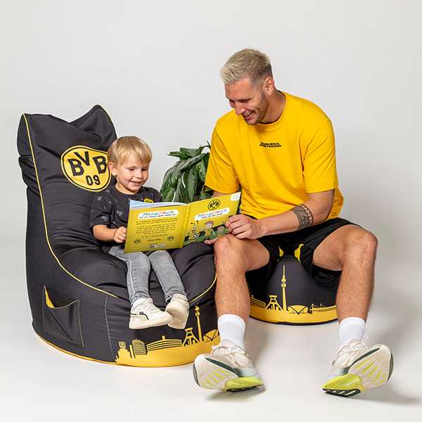 Ein Erwachsener und ein Kind sitzen auf schwarzgelben Sitzsäcken mit BVB-Emblem und lesen gemeinsam das BVB Mitmachbuch, Frontalansicht.