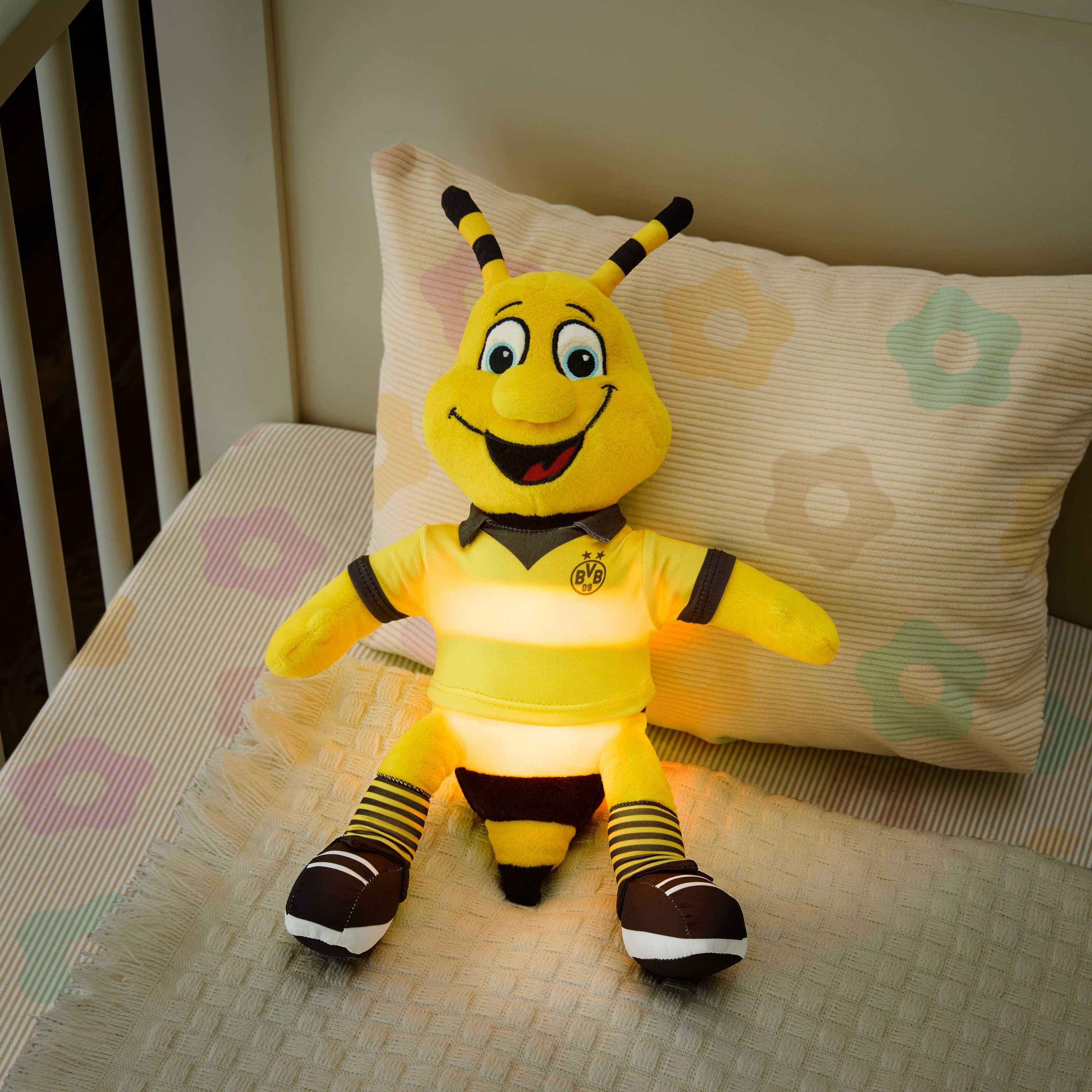 Vorderansicht des BVB Nachtlicht EMMA als Plüschfigur im schwarzgelben Trikot mit leuchtendem Bauch und sichtbarem BVB-Emblem, auf einem Kinderbett.