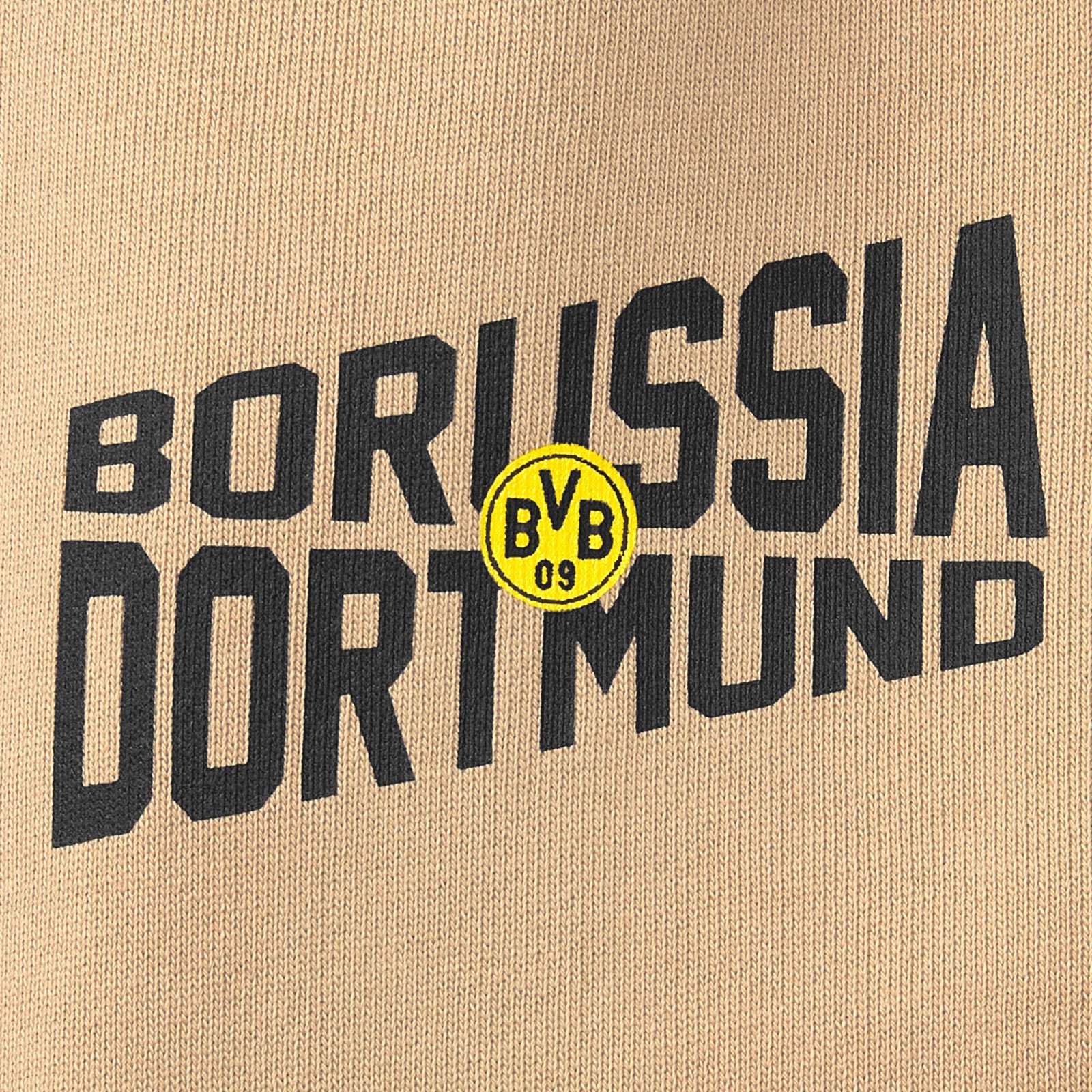 Detailansicht der BVB Sweatpants beige Kinder mit großem schwarzen "Borussia Dortmund"-Schriftzug und gelbem BVB-Emblem auf beigem Stoff.
