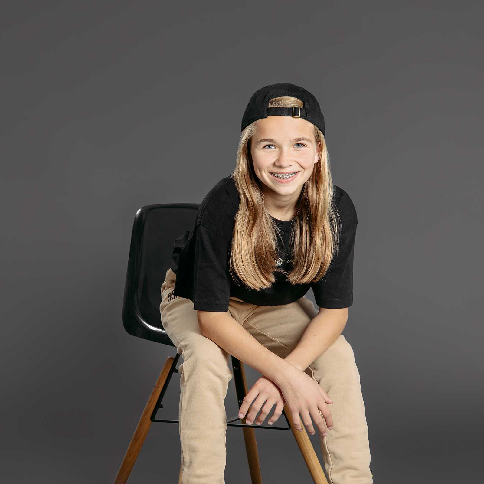 Ein Kind trägt in der Frontansicht das BVB T-shirt oversized Kinder in schwarz auf einem Stuhl, lächelt und trägt eine schwarze Cap. Das BVB-Emblem ist dezent sichtbar.