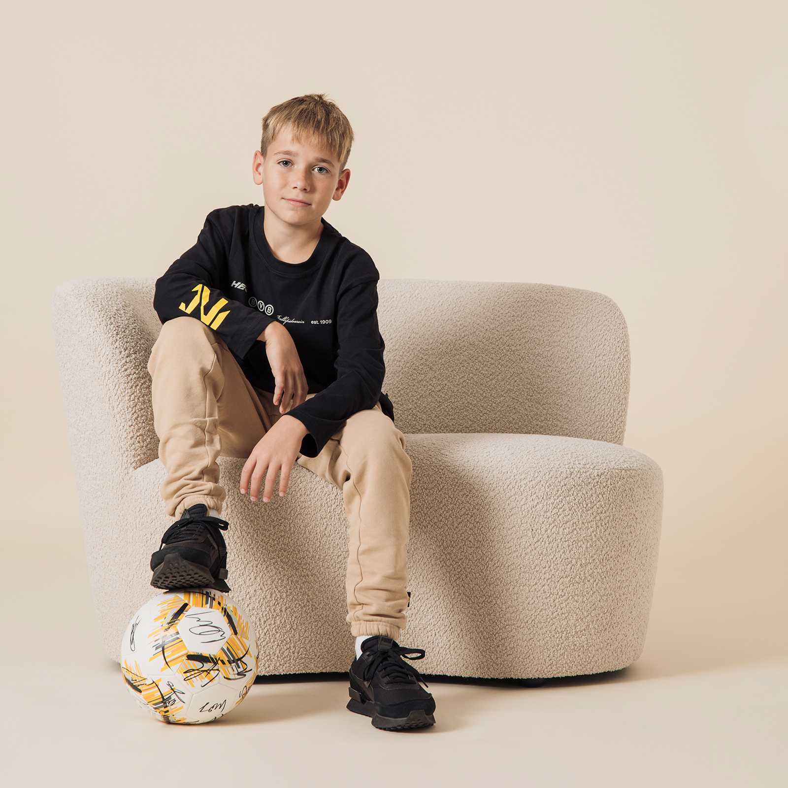 Ein Junge trägt das schwarzgelbe BVB Longsleeve oversized Kinder, sitzt auf einem Sofa und hat einen Fußball am Fuß. Vorderansicht des Produkts.