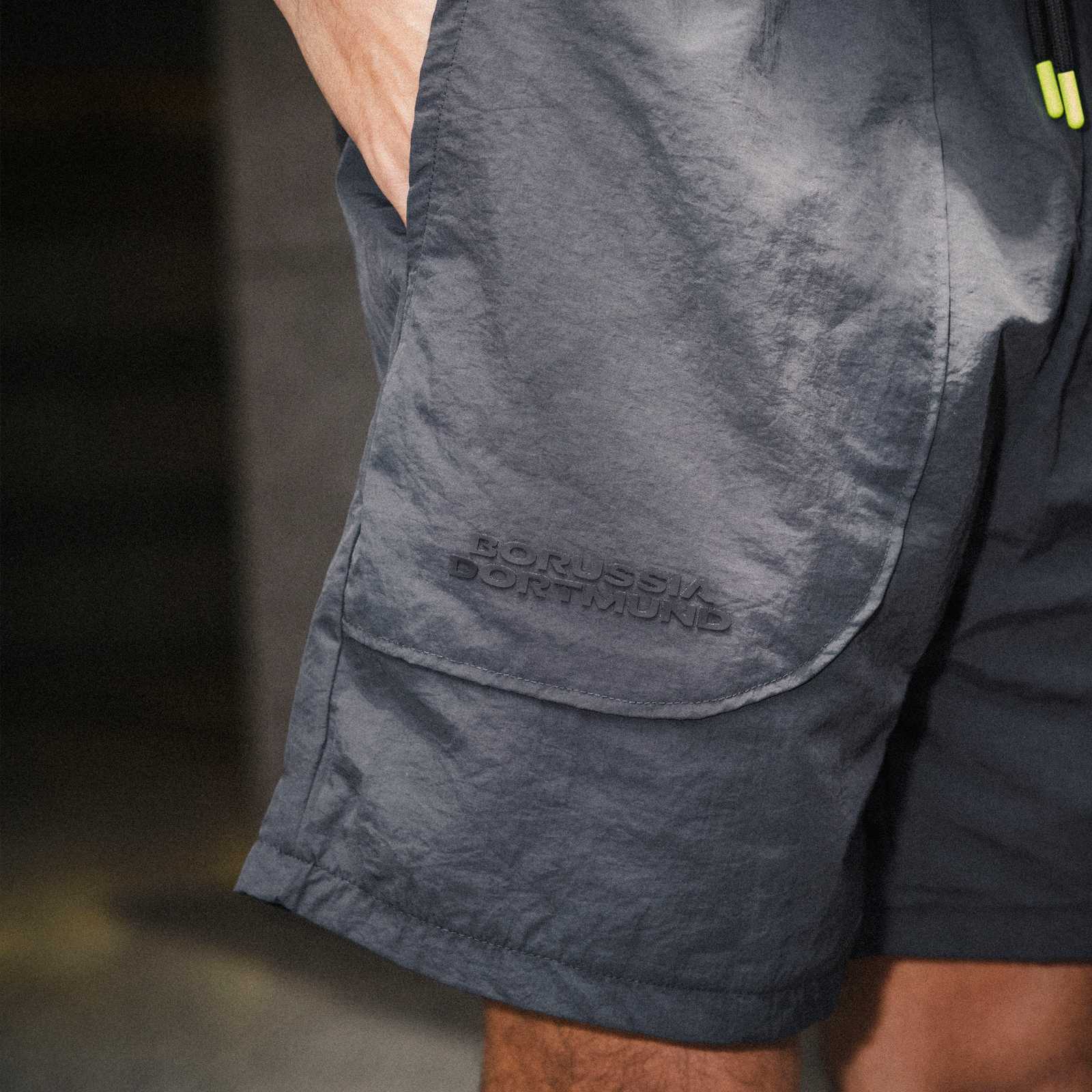 Detailansicht der BVB Underground Woven Shorts in Dunkelgrau mit geprägtem "Borussia Dortmund"-Schriftzug auf der Seitentasche. Person trägt die Shorts.