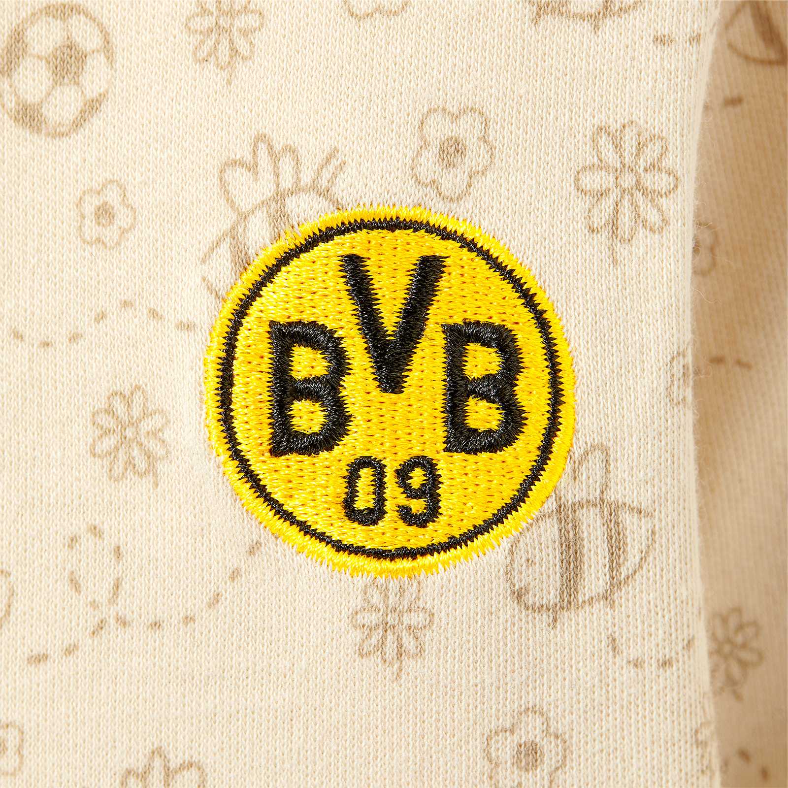Detailansicht des BVB Body 2er-Set Baby mit gesticktem BVB-Emblem auf beigem Stoff mit dezentem Blumen- und Fußballmuster.
