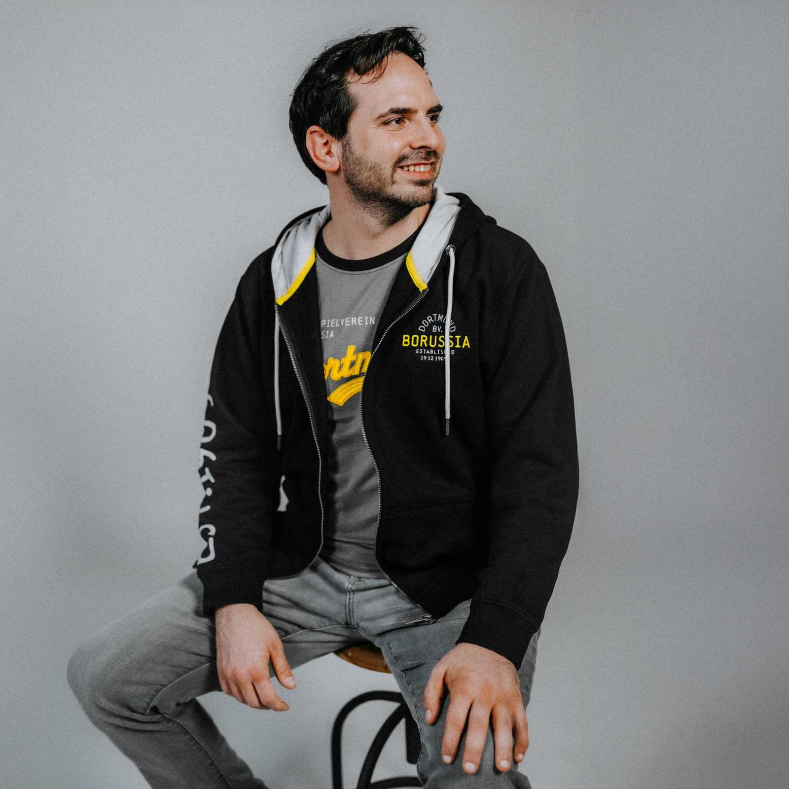 Ein Mann trägt die schwarzgelbe BVB Explorer Sweatjacke in der Vorderansicht, mit auffälligem Borussia Dortmund Schriftzug auf der Brust und Kapuze mit gelbem Innenfutter.
