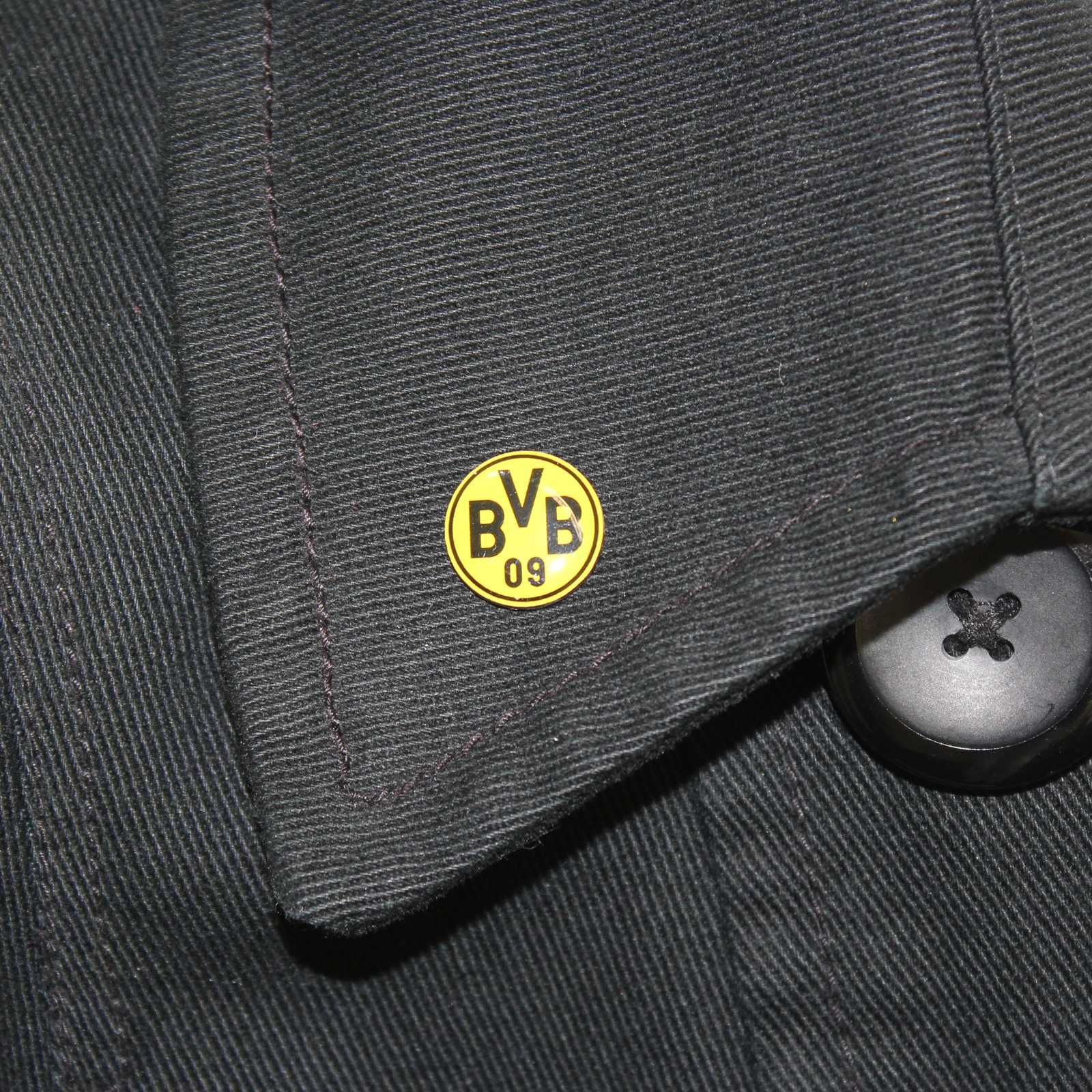 Detailansicht des BVB Pin Emblem gelb, rund und mit schwarzem BVB-Emblem, an einem schwarzen Stoffkragen befestigt.