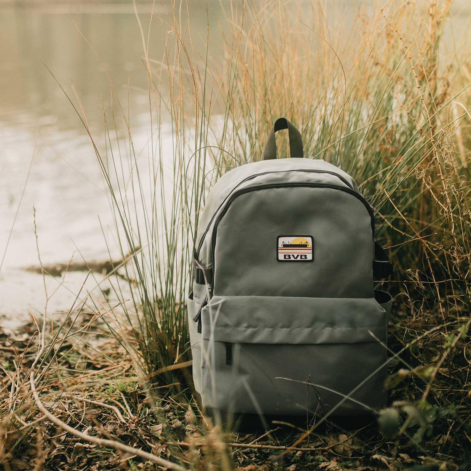 Vorderansicht des BVB Sunrise Rucksacks in Grau mit aufgenähtem BVB-Emblem und Sonnenaufgang-Design, fotografiert in der Natur.