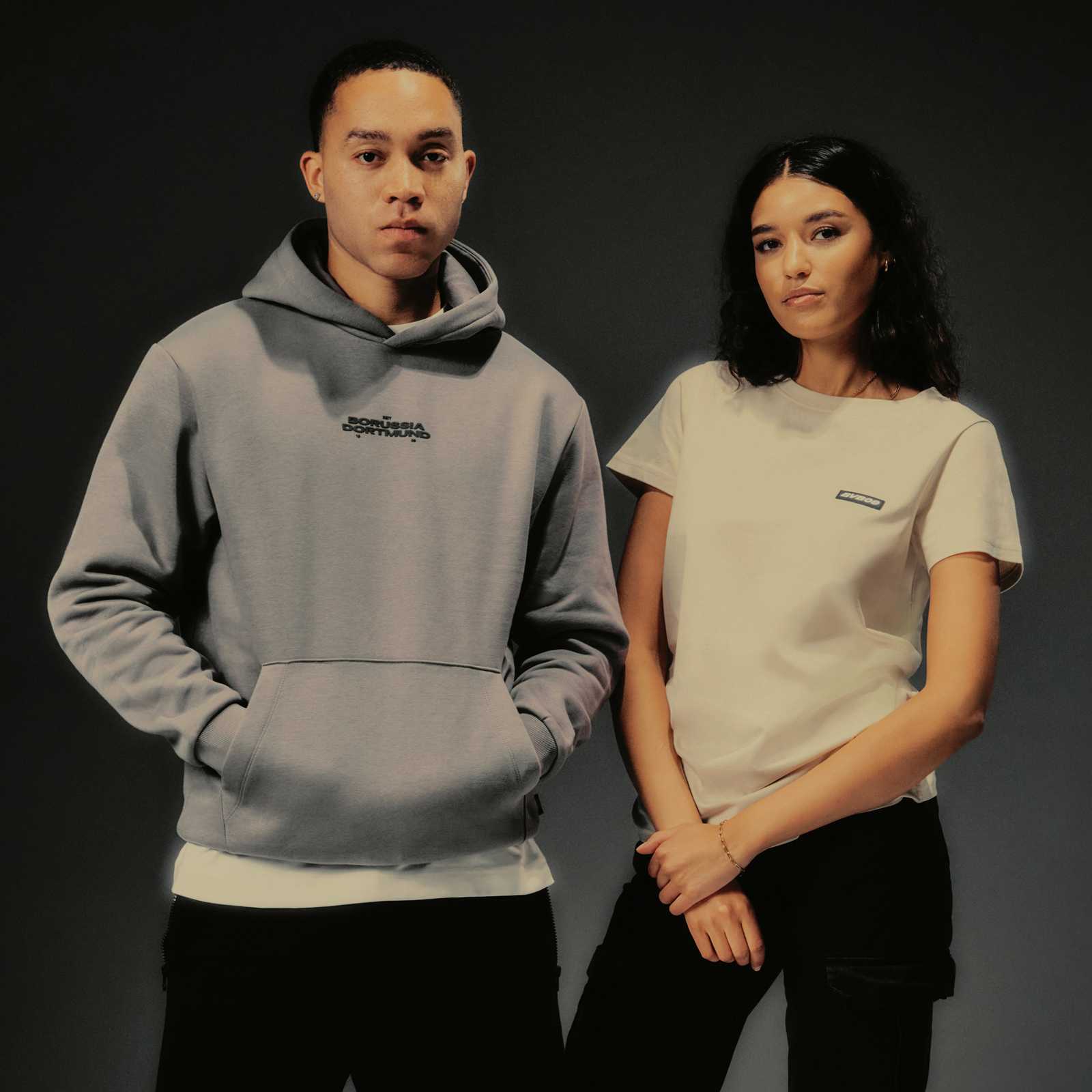 Ein männliches Model trägt den Essentials Hoodie, ein weibliches Model das beige Essentials T-Shirt