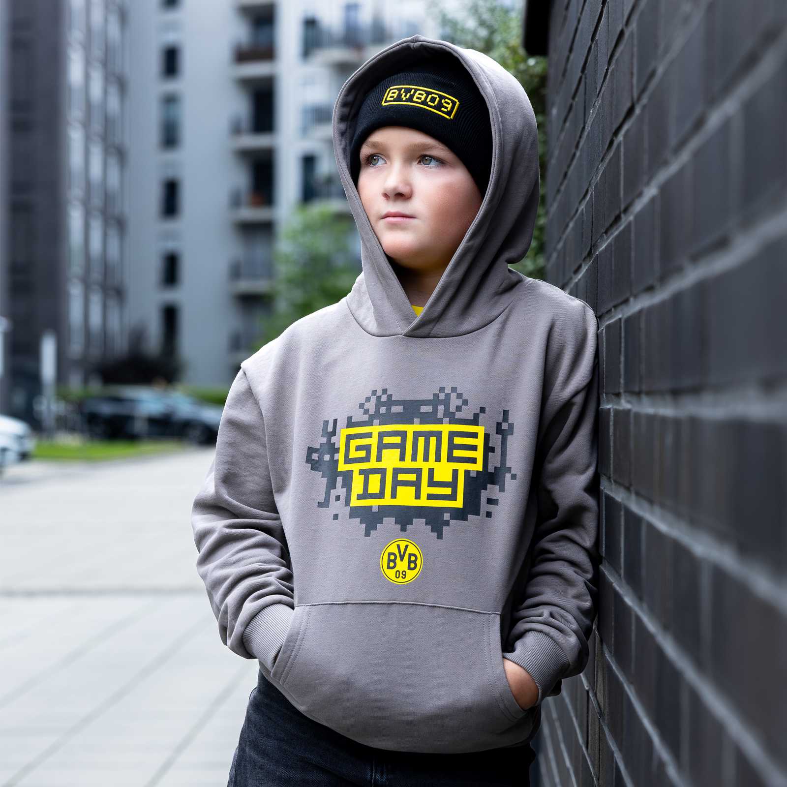Kleiner junge lehnt an einer Mauer und trägt den grauen BVB Hoodie mit dem Game Day Print. Passend dazu trägt der junge eine schwarze BVB Mütze und die Kapuze des Hoodies drüber.