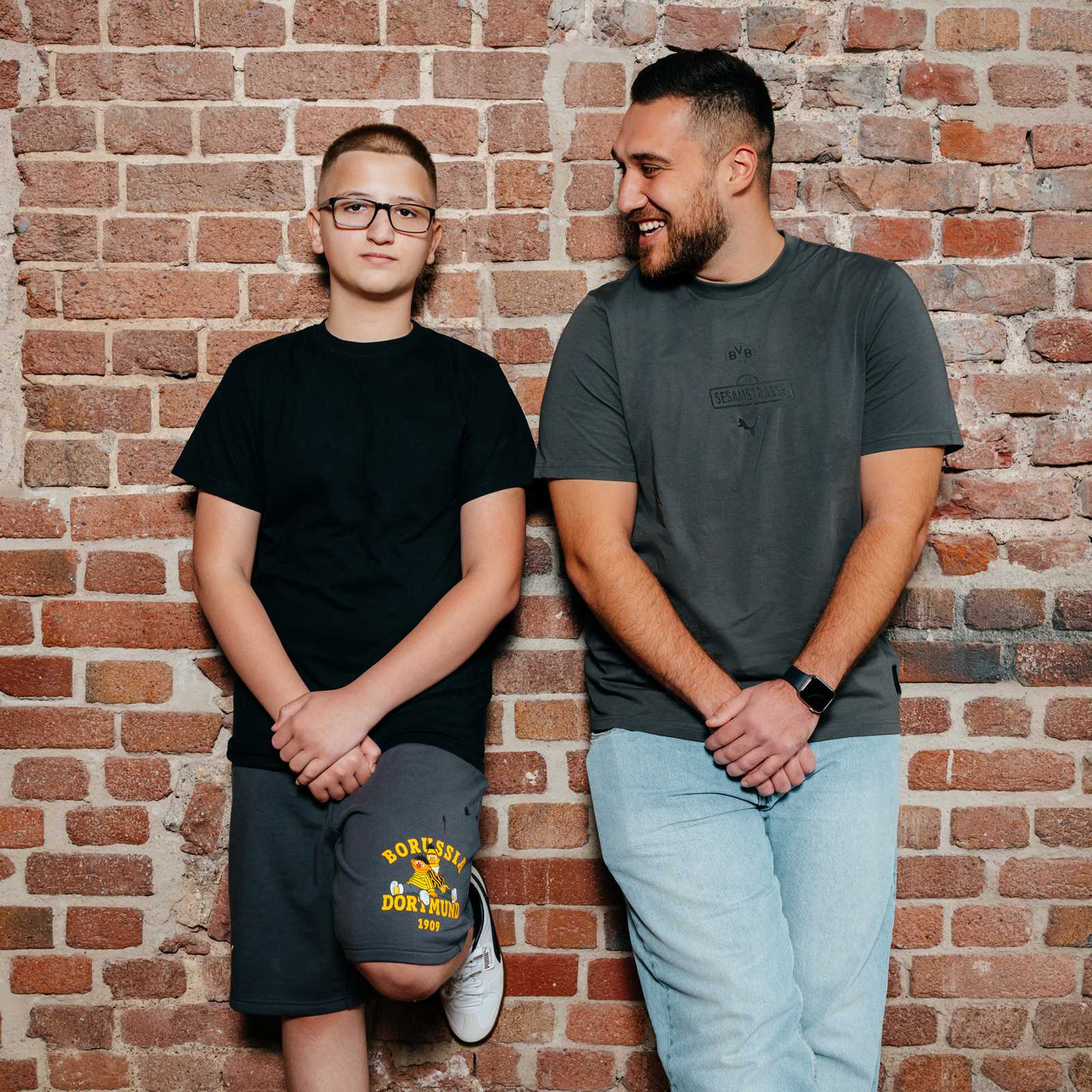 Ein männliches junges Model lehnt an einer Wand neben einem älteren Model mit Bart und trägt die grauen Shorts.