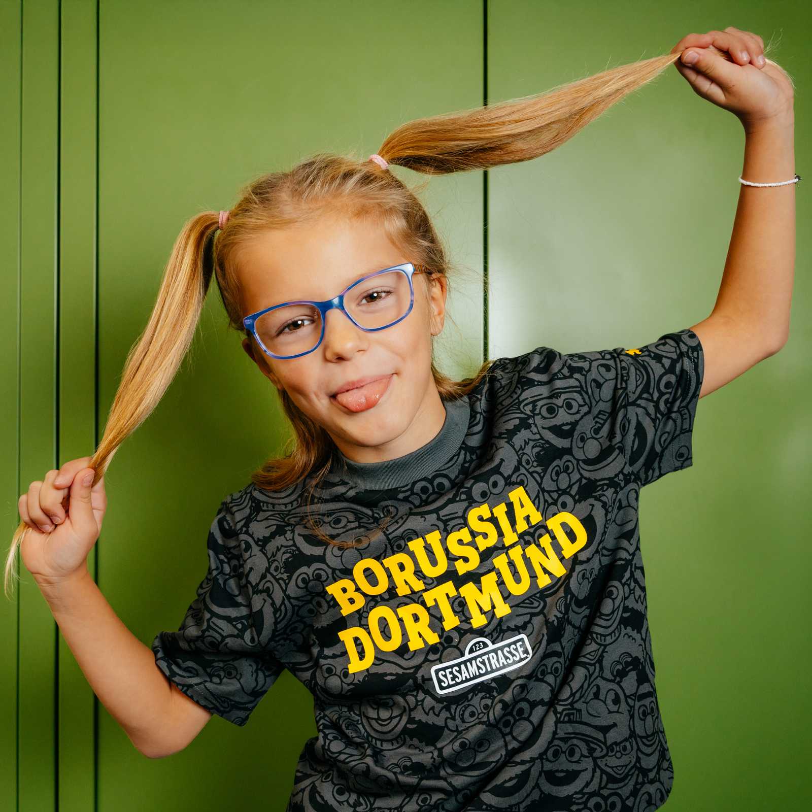 Ein blondes Mädchen mit Brille trägt das grau-schwarze T-Shirt und streckt die Zunge raus.