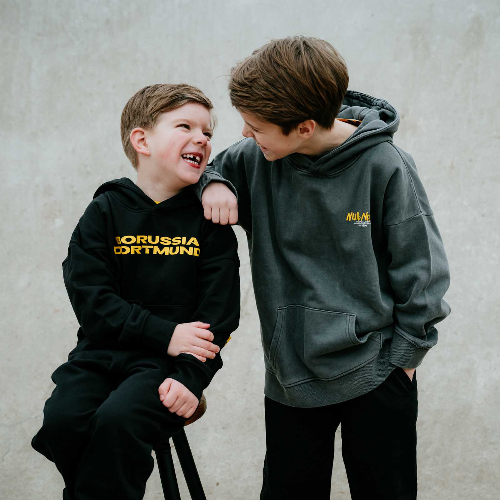 Zwei Jungen stehen nebeneinander und tragen BVB-Pulli. Der linke Junge trägt den schwarzen Hoodie mit Borussia Dortmund Print.