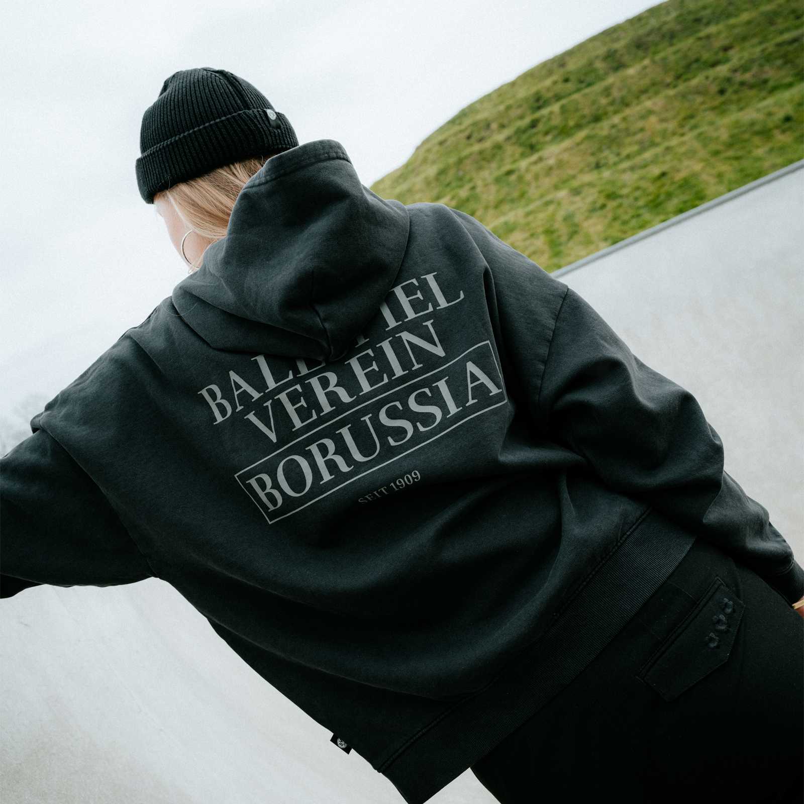 Ein weibliches blondes Model stützt sich mit einer Hand am Rand einer Skate-Bahn ab und trägt den Hoodie. Man sieht die Rückseite.