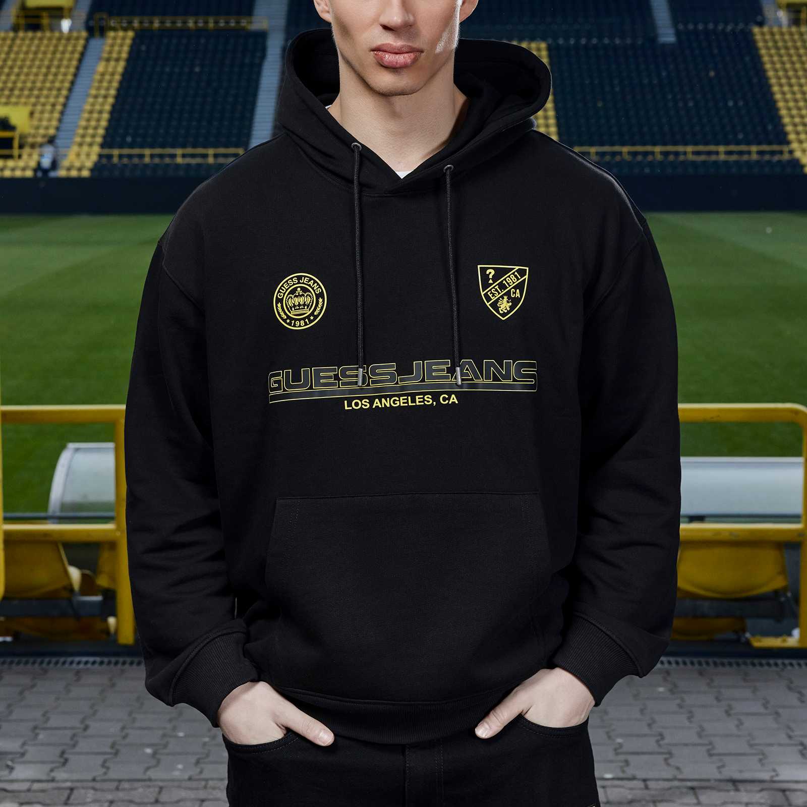 Junges männliches Model steht im Stadion und trägt den schwarzen BVB-Hoodie mit gelben Prints.