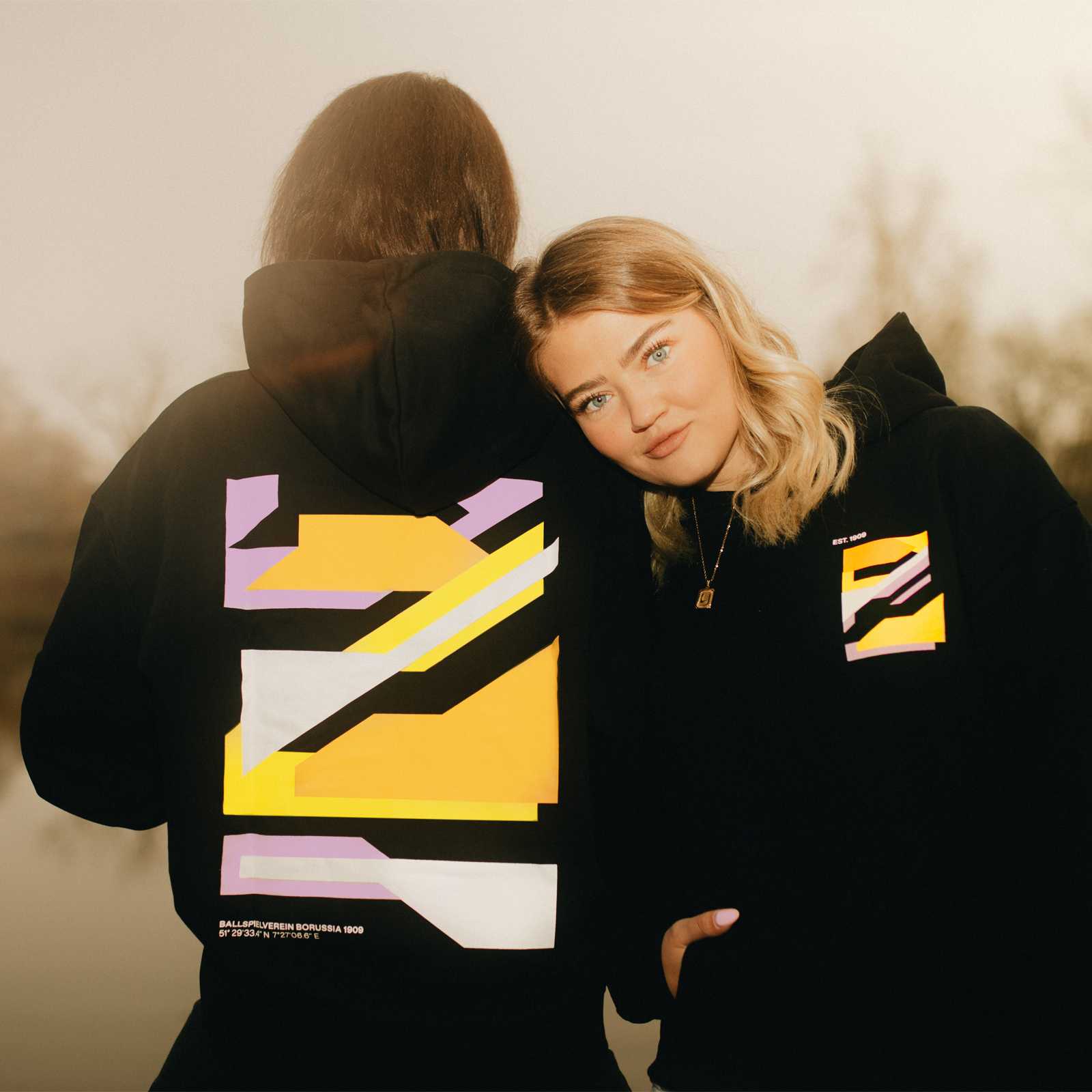 Zwei junge Frauen stehen nebeneinander und tragen den schwatzen BVB-Hoodie. Eine steht mit dem Rücken zur Kamera und die andere schaut direkt in die Kamera.